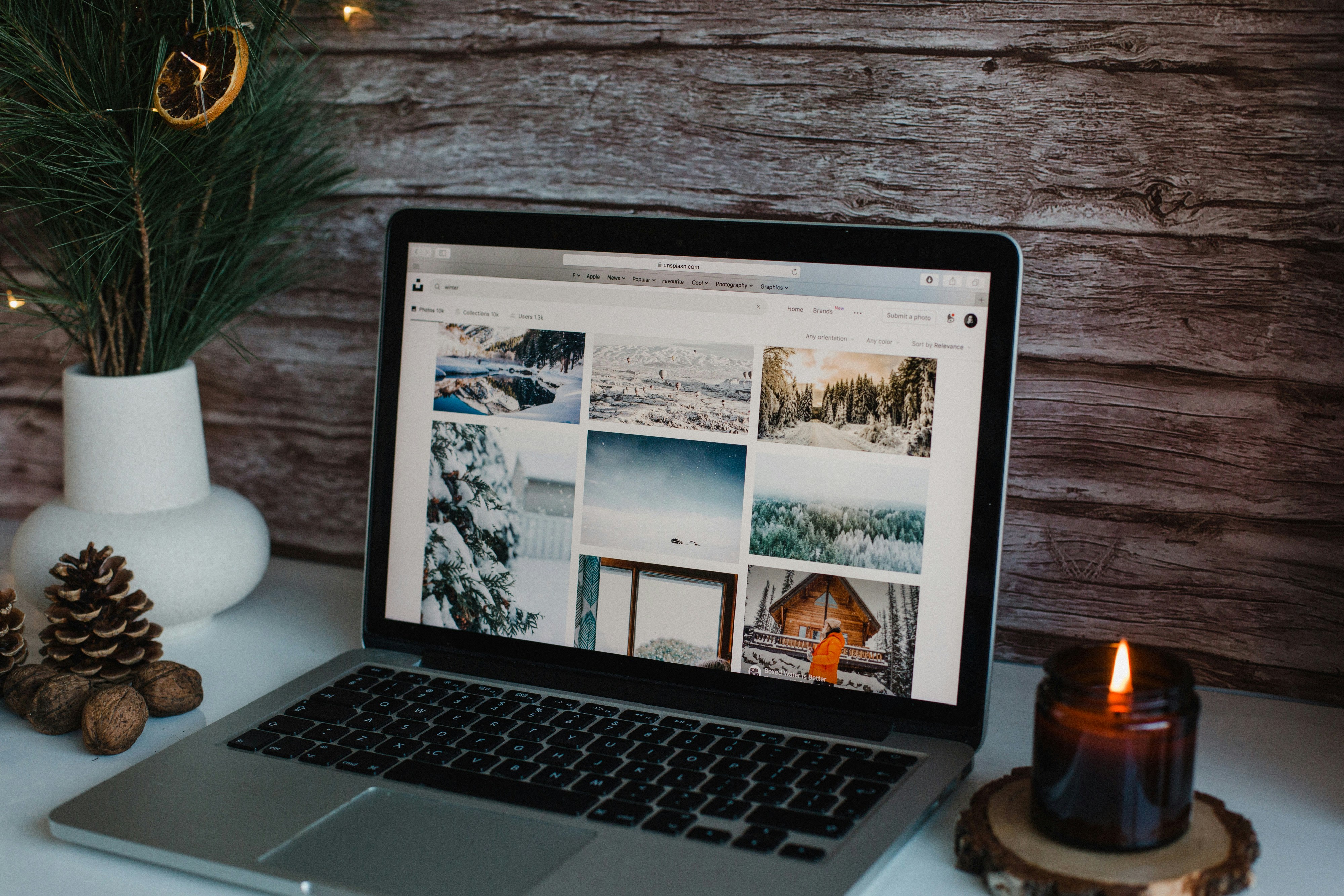 Foto eines Laptops der winterliche Landschaften zeigt. Um den Laptop sind Tannenzweige, Tannenzapfen und eine Kerze angeordnet.