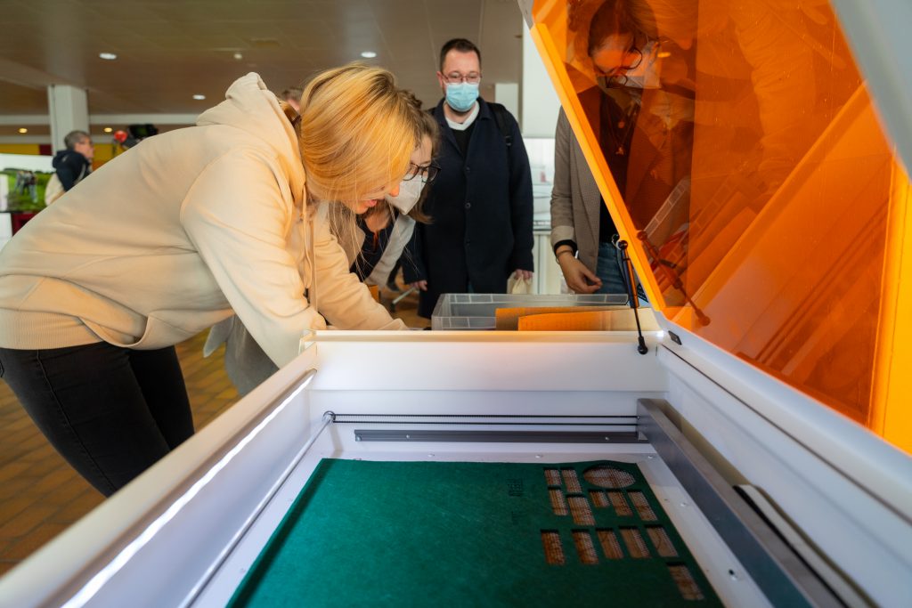 geöffneter Lasercutter im Vordergrund, mehrere Personen im Hintergrund
