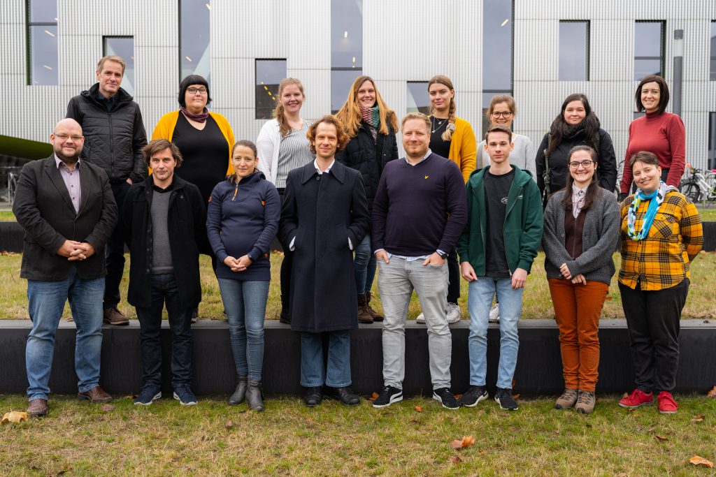 16 Teammitglieder im Projekt UOS.DLL. Sie stehen zweireihig vor einem grauen Universitätsgebäude mit herbstlichen Klamotten an.