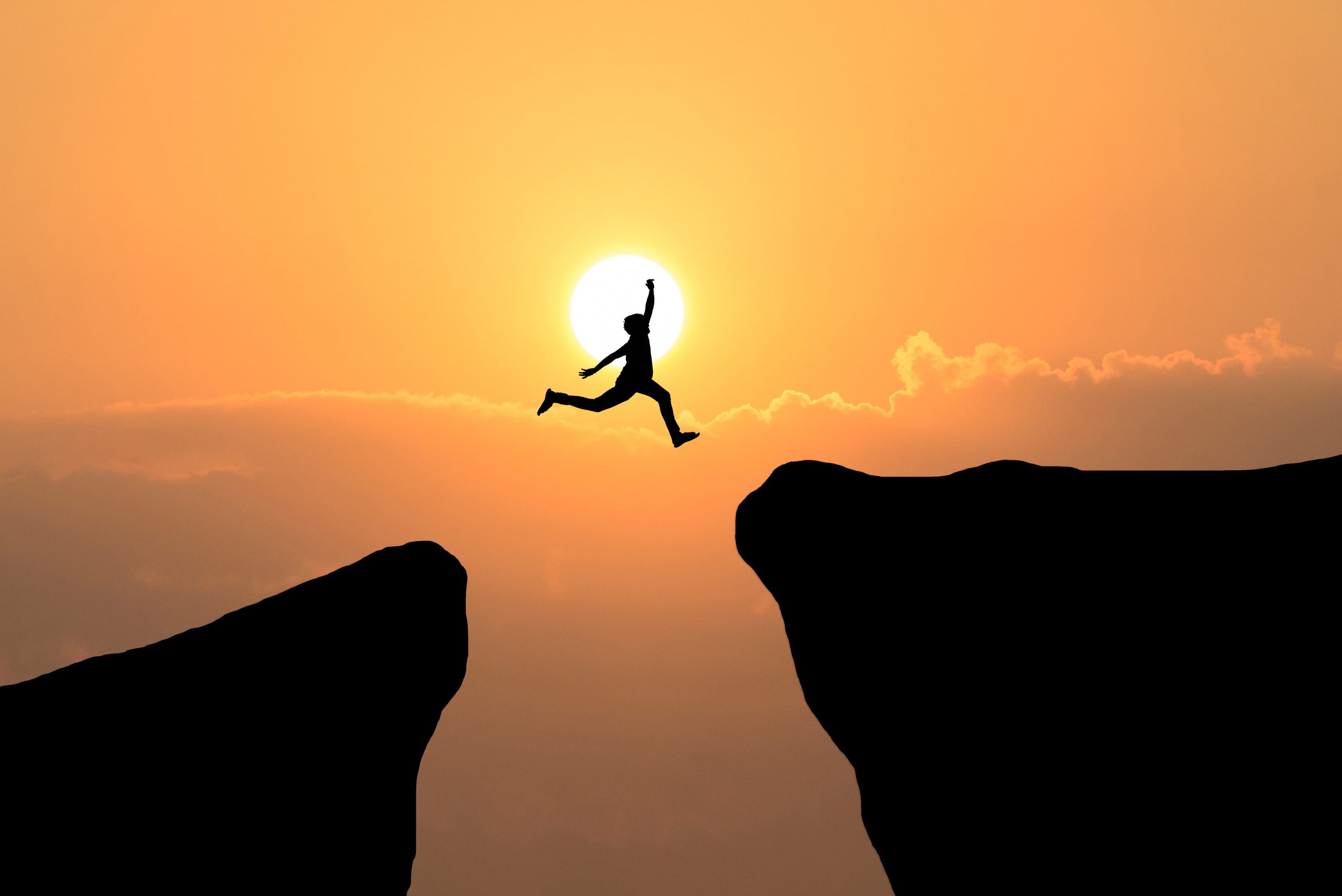 Eine Person springt von dem einen zum anderen Felsen. Im Hintergrund ist die Sonne zu sehen.