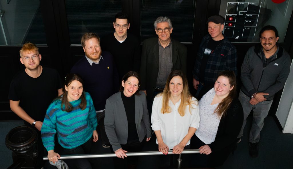 10 Teammitglieder im Projekt SOUVERÄN. Sie stehen zweireihig in einem abgedunkelten Raum und gucken von unten hoch in die Kamera.