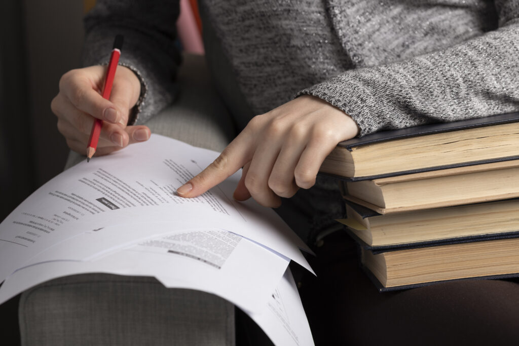 Nahaufnahme von einer Person, die mit einer Hand auf ein mit Text bedrucktes Papier zeigt und in der anderen Hand einen Stift hält. Daneben liegt ein Stapel Bücher.
