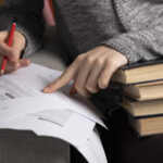 Nahaufnahme von einer Person, die mit einer Hand auf ein mit Text bedrucktes Papier zeigt und in der anderen Hand einen Stift hält. Daneben liegt ein Stapel Bücher.
